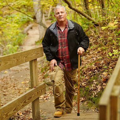 Man standing on a wooden path in a forest, holding a walking stick.