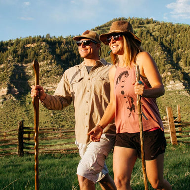 Two people holding hands with hiking sticks in a scenic outdoor setting with mountains and a fence.