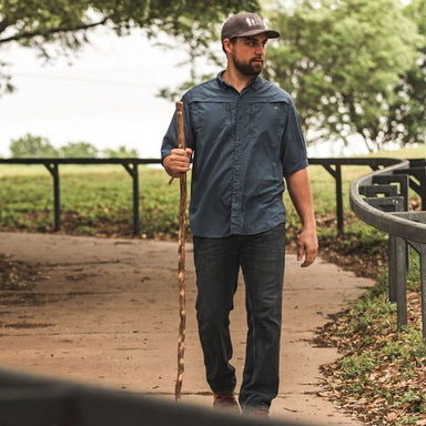 Man walking outdoors holding a walking stick, wearing a blue shirt and cap.