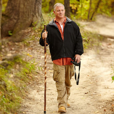 Man walking on a forest path with a walking stick and camera, wearing a black jacket and beige pants.