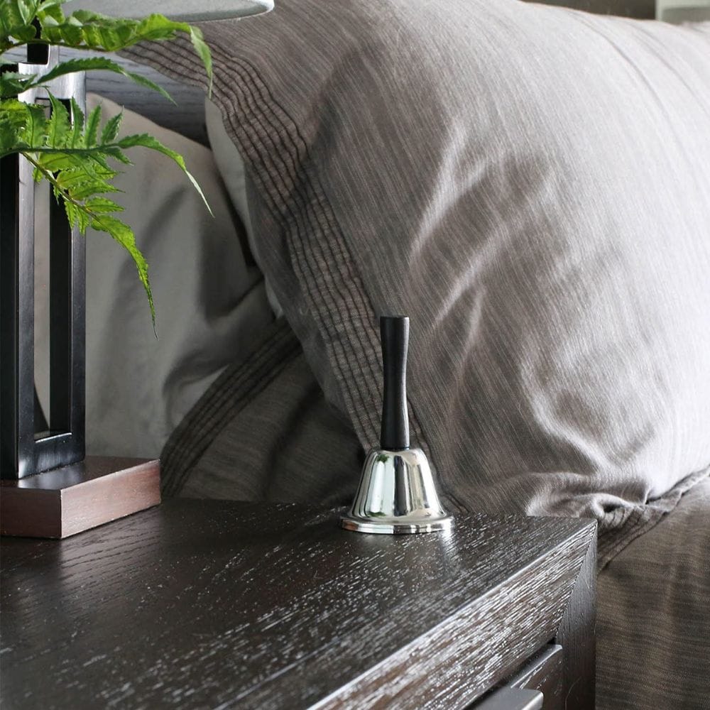 Silver bell on a wooden nightstand next to a bed with gray bedding