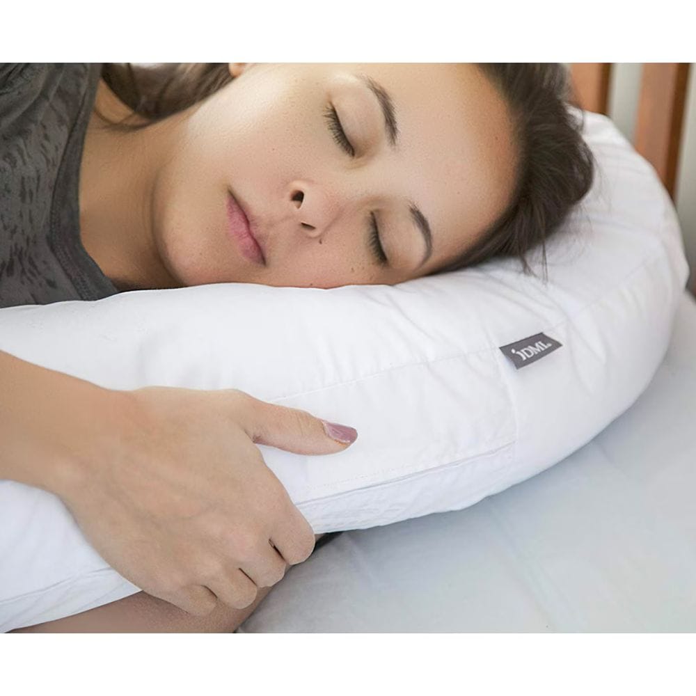 A woman sleeping peacefully on a white U-shaped cervical pillow, with her hand resting on the front. The pillow features a small black brand tag reading 'XCEL'.