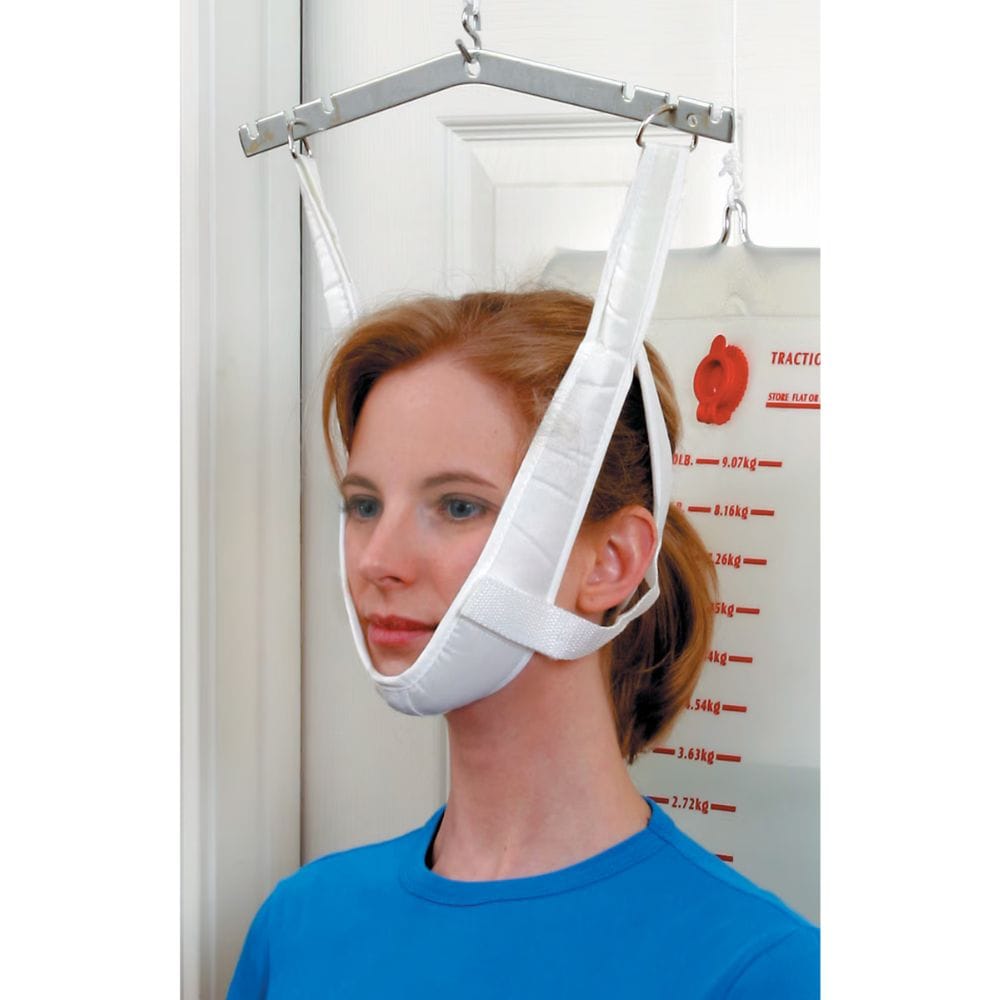 A woman wearing a white chin-lower support harness suspended from a metal frame, shown indoors against a white door and weight-measuring wall.