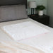 White heating pad on a bed with a nightstand and lamp in the background