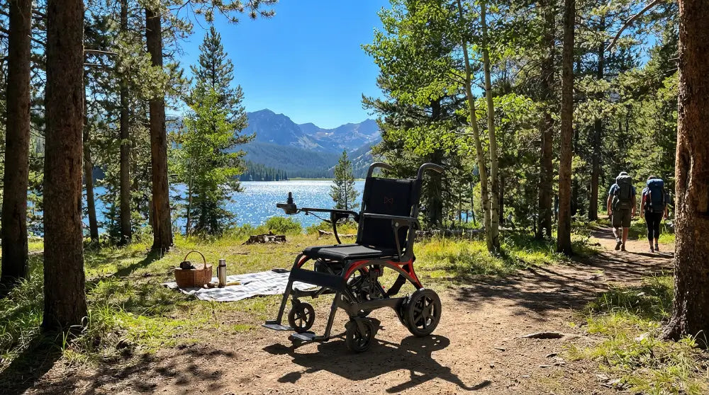 Monarch Mobility Ezi-Fold Carbon power wheelchair with orange frame accents on a wooded lakeside trail with mountains in the distance.