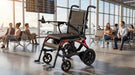 Monarch Mobility Ezi-Fold Carbon power wheelchair with orange frame accents displayed in an airport terminal, highlighting its travel-friendly design.