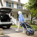 Woman pushing a stroller next to an open car trunk in front of a house.