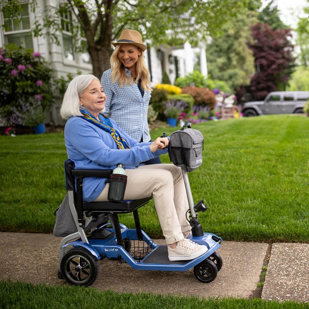 Woman using a mobility scooter with another woman standing behind her on a sidewalk.