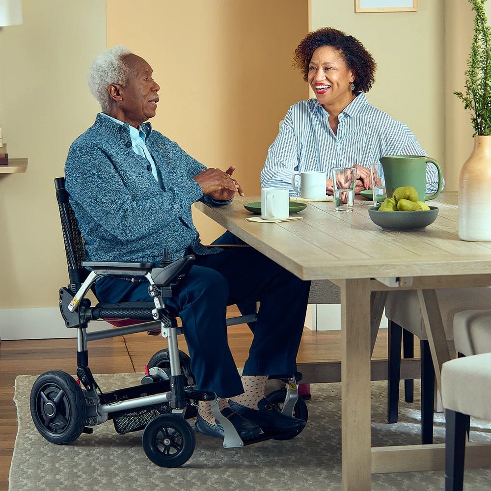 Man in a wheelchair and woman sitting at a dining table, smiling and engaged in conversation.