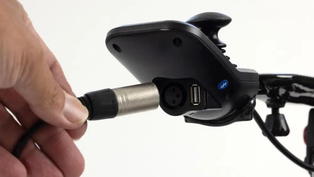 A close-up of a hand plugging a black and silver cable into a port on a black electronic device mounted on a bicycle handlebar.