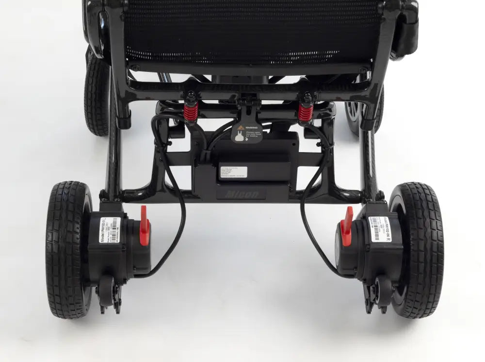 Close-up rear view of a black electric wheelchair frame with four wheels, black mesh seat, red suspension springs, and battery units mounted on the rear wheels against a white studio background.