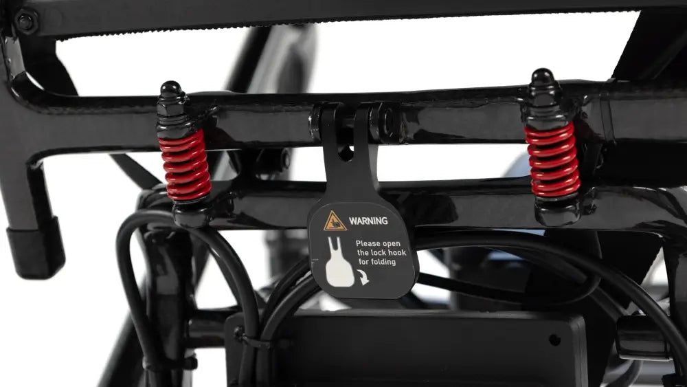 Close-up of a black metal frame component with two red coil springs and a hanging black tag that reads WARNING with a rabbit icon and folding instruction.