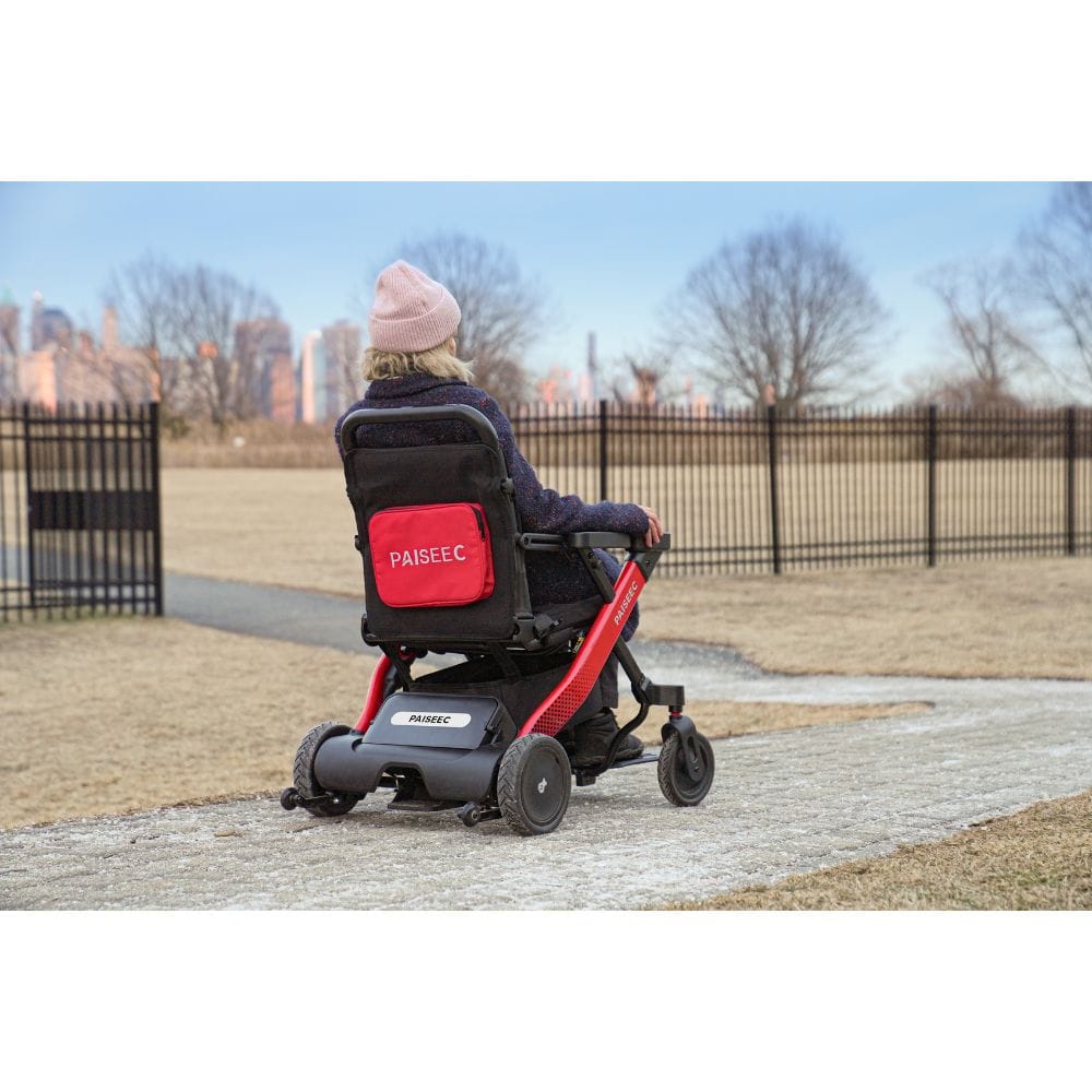 Person using a PAISEEC electric wheelchair in an outdoor setting with a cityscape in the background.