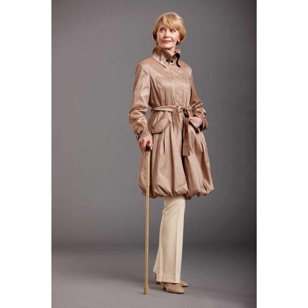 Woman wearing a beige trench coat and holding a cane against a gray background