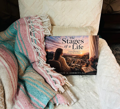 The Stages of a Life by Julie A. Corbett is displayed on a soft white chair beside a colorful woven blanket. The cover features a woman reflecting on the memories of her life against a peaceful sunset landscape.