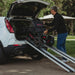 Man loading a mobility scooter onto a car using a ramp in an outdoor setting.