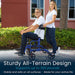 Man using a blue all-terrain walker with a woman assisting him outdoors.