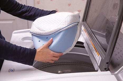 A person places the white-covered Contour Health Legacy Leg Pillow into a washing machine.