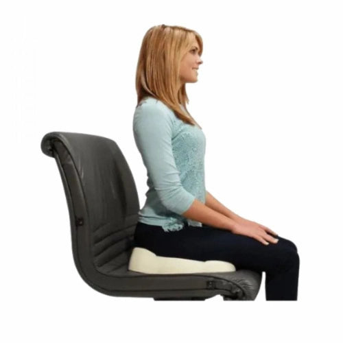A woman in a light blue top and dark pants sits in profile on a black ergonomic office chair with a cream-colored lumbar support cushion, shown against a plain white background.