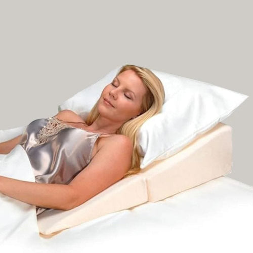 A woman reclining on a white pillow and cream-colored wedge bed support pad against a plain light gray background.